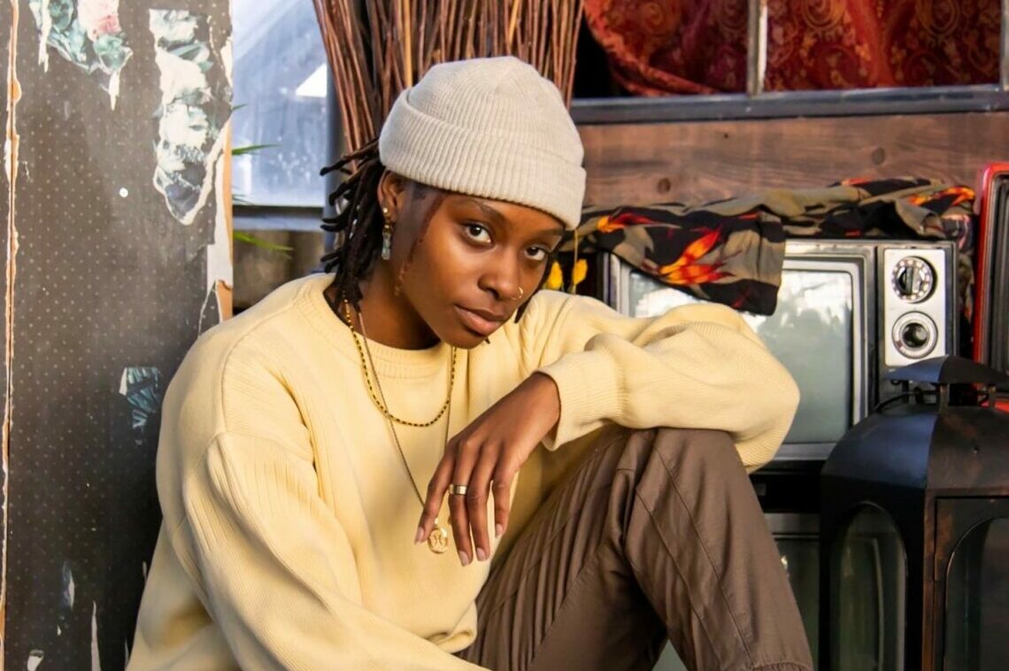 DJ Shannyn Hill sitting with her legs bent, wearing a yellow crewneck and a grey beanie.