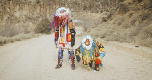 Two people are beside each other in a desert, one standing and one kneeling. They are looking down at the ground, and are wearing colourful Indigenous clothing and headwear.