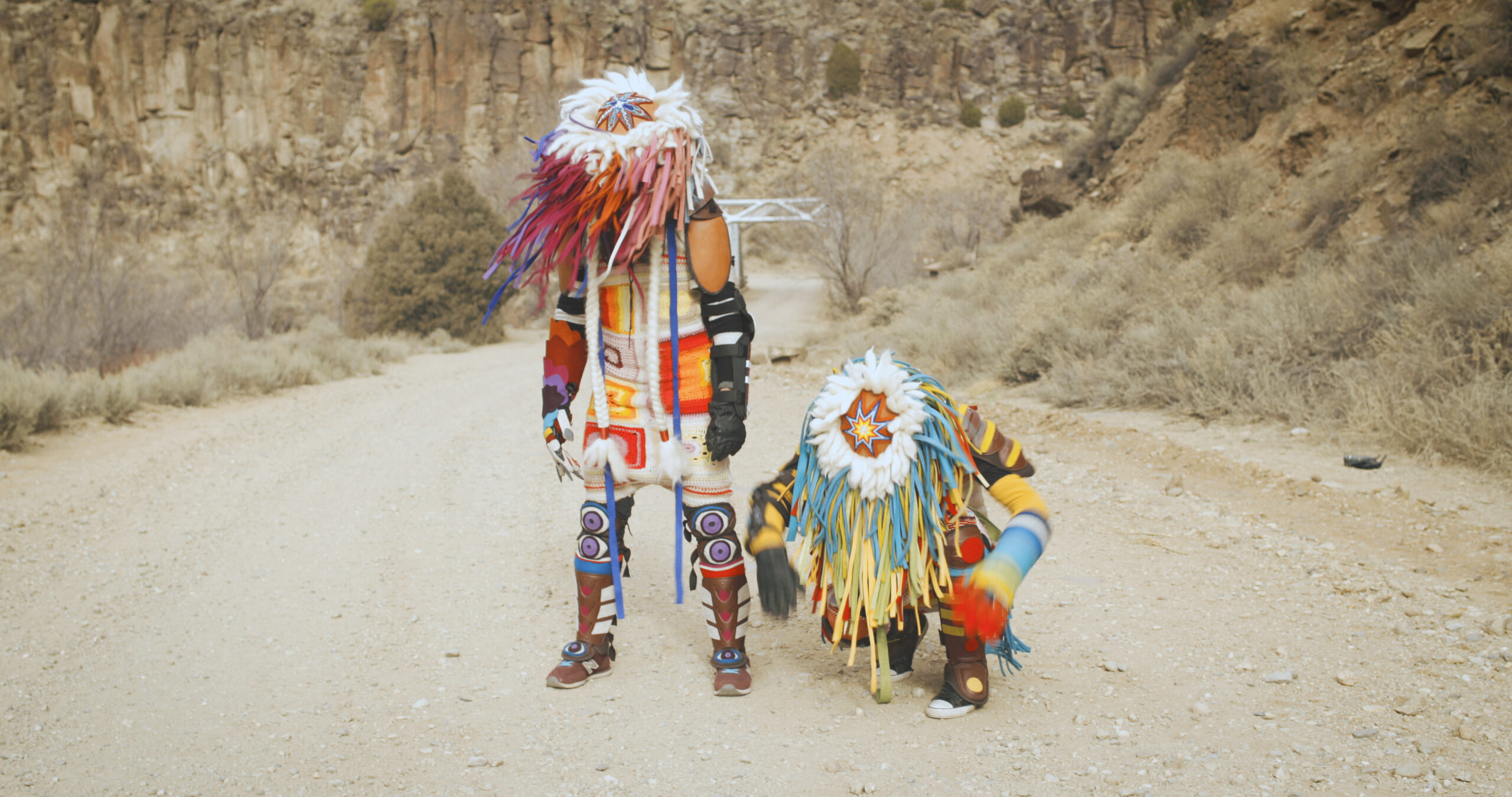 Two people are beside each other in a desert, one standing and one kneeling. They are looking down at the ground, and are wearing colourful Indigenous clothing and headwear.