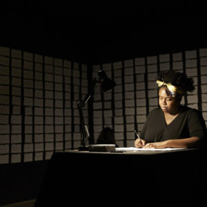 A woman sits in a dark room writing in a notebook, only illuminated by a lamp on a desk.