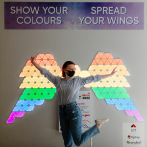 A woman posing in front of a wall that includes a pair of wings made from flat LED lights. The lights are lit up in the colours of a rainbow flag.