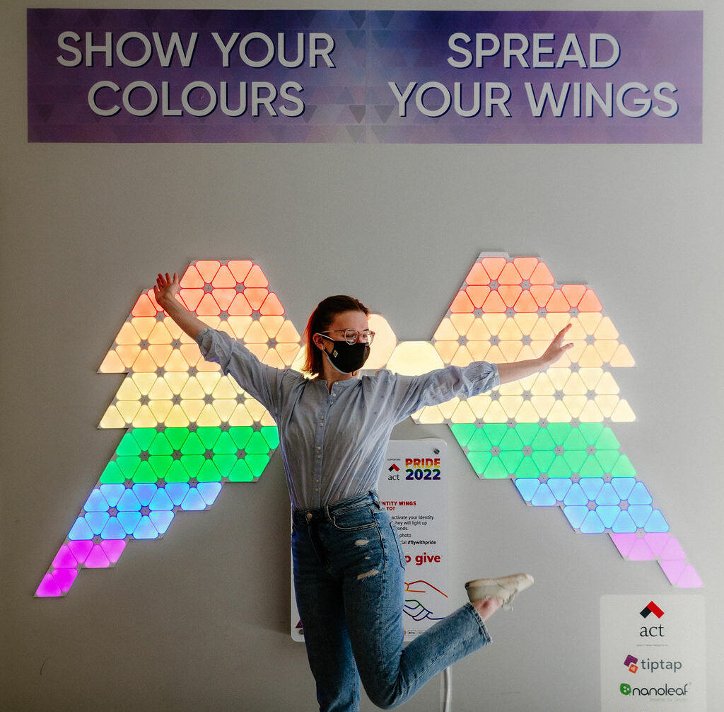 A woman posing in front of a wall that includes a pair of wings made from flat LED lights. The lights are lit up in the colours of a rainbow flag.