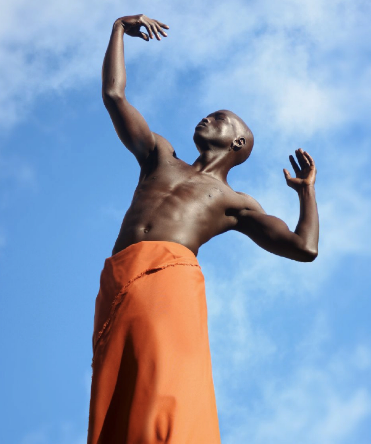Photograph of a richly-pigmented Black figure whose lower body is wrapped in orange cloth, posing against the backdrop of a blue sky.