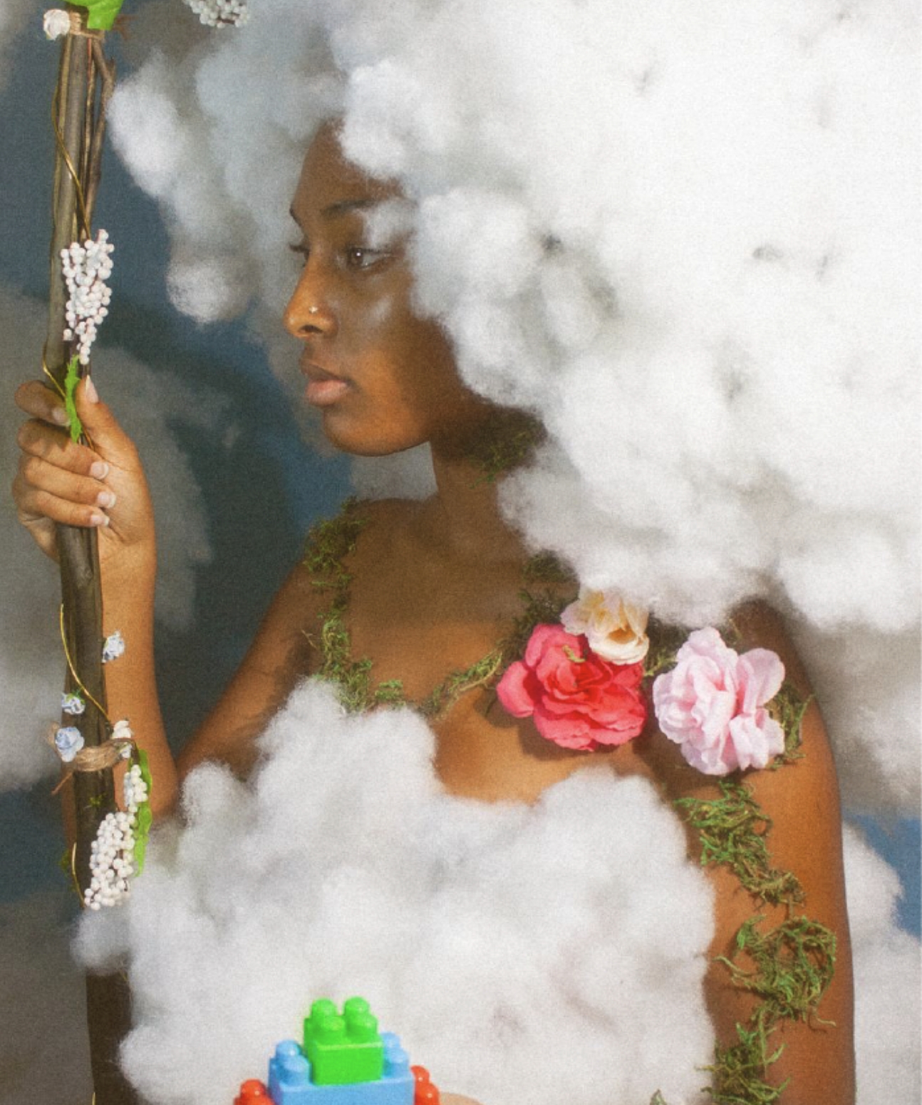 Photograph of a Black woman whose clothes and hair are made of clouds, holding a wooden stick in one hand, and multicoloured building blocks in the other. Pink flowers and green moss are growing across her arms and shoulders